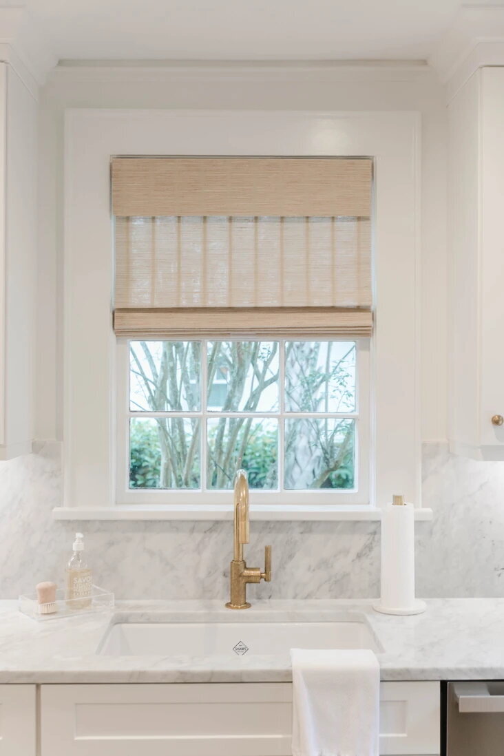 Coastal kitchen with marble countertops and backsplash, brass fixtures and woven window treatments.