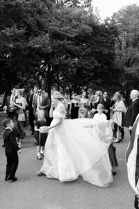 Black and white image of a bride walking to the ceremony location with her wedding dress train being fluffed and blown up by the wind in this stunning editorial profile view
