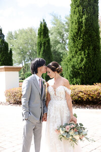 Groom in grey suit kissing the forehead of his bride wearing a lace dress as they stand outdoors