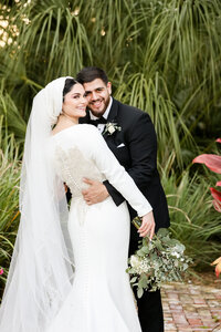 Bride and groom hugging and looking at the camera in an outdoor setting surrounded by palms and greenery