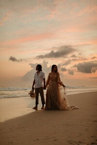 Bride and groom holding hands at sunset on beach with ocean behind them