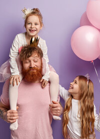 Papa en rose tenant sa fille en tenue de princesse, souriante, sur ses épaules. A côté son autre fille tenant des ballons. Fête anniversaire organisée par une agence évènementielle.