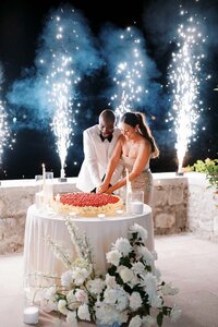 Bride and groom cutting a wedding cake surrounded by sparklers and candlelight during an evening celebration by the sea
