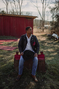 Man in a leather jacket  poses next to a red barn with a black cowboy hat and cowboy boots.