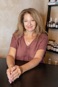 Cyndi Stewart sits at a table in her holistic practice room, smiling warmly with shelves of essential oils behind her.
