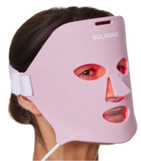 A woman wears a light pink LED therapy face mask by Solawave, which wraps around the face and has cutouts for the eyes, nose, and mouth. The design is sleek and futuristic, used for skincare and light therapy treatments.