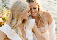 Two brides in white dresses share a close and affectionate moment, with one nestling her head on the other’s shoulder