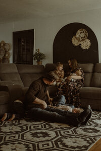 A family of 5 sit in their living room, welcoming the newest addition. Dad is on the floor playing with the boys while mom sits on the couch with her girls...her oldest and youngest.