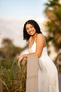 Stunning senior photos in Central Florida at the iconic Cocoa Beach Pier. Senior girl is wearing pink shirt, blue jean shorts, and has brown curly hair. She is positioned seated with hands crossed over one knee. 