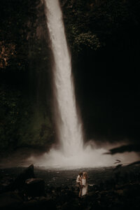 A man and a woman stand at the foot of a tall narrow waterfall.