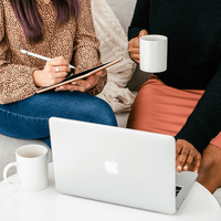 Two women sitting on a couch, one writing on a tablet, the other holding a coffee mug. An open laptop is on a table, creating a collaborative mood.