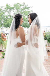 Two women in white wedding dresses and veils stand facing each other, holding hands and smiling. They are outdoors on a brick pathway, surrounded by colorful flowers and greenery.