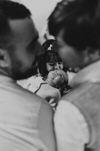 Isaline et son chéri de dos prêts à s'embrasser et dans l'encadrure de leurs visage, leur fille portant bébé lors d'un shooting photo naissance en Vendée.