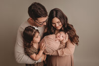 Family of 4 portrait standing up wearing neutral pinks, browns and creams, with mom holding newborn  baby girl and mom, dad and 2 year old sister are snuggled in close looking at baby and smiling at her
