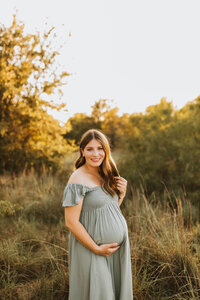 brunette pregnant mother in a sage green dress in a field at sunset