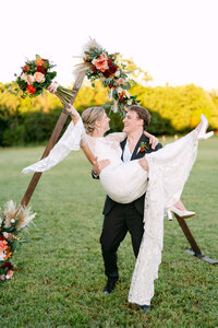 A groom picking up his wife and she's cheering with her hands up and she has her bouquet on her hand..