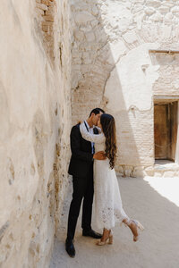 Couple kissing in an intimate moment against the warm stone walls of a historic mission building.