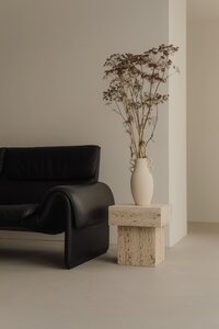 A modern minimalist room featuring a black leather sofa, a beige marble side table, and a large clay vase with dried branches. The neutral tones create a calm, elegant aesthetic.
