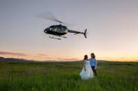 Bride and groom stand in a wildflower-filled Montana field at sunset as a black helicopter hovers above, capturing the excitement of their adventure elopement. The bride wears a flowing white wedding dress, and the groom, dressed in a blue shirt and suspenders, smiles as they soak in the moment.