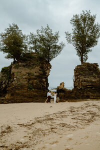 M & J facing one another holding each other tight while standing on a rock surrounded by ocean and rock formations on each side of them
