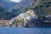 A picturesque coastal village with white buildings nestled on a hillside. The village overlooks a deep blue sea, surrounded by lush green mountains. Stone arches and terraced fields are visible, adding to the scenic landscape.