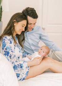 Portrait of a woman in a blue floral dress and a man in a blue shirt and khaki pants sitting smiling over a baby boy on a bed