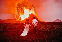 Bride & groom holding hands walking over lava rock toward erupting volcano!