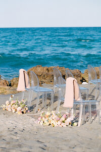 Two Wedding chairs ringed with flowers sit alone at the beach.