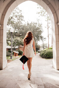 Brunette walks away through arched stone holding a graduation cap in the left hand. 