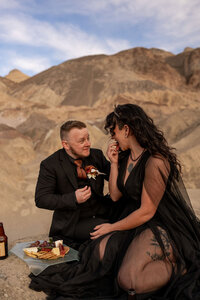 A bride in a black wedding dress standing against the vibrant, rainbow-colored hills of Artist's Palette in Death Valley, a symbol of uniqueness and love