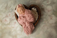 newborn baby girl in heart bowl with flower backdrop and pink swaddle