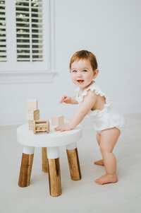 Baby girl in white romper stands at stool with wooden toys and smiles during baby photography session in Raleigh NC