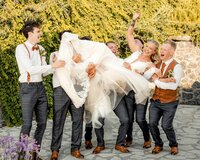 A joyful group of groomsmen lifting a laughing bride in her wedding dress, surrounded by greenery and stone walls, captured during a candid wedding party moment by a UK wedding photographer.