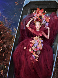 A captivating outdoor portrait of a young girl lying elegantly in a rustic metal boat, surrounded by vibrant autumn foliage. She wears a stunning deep red tulle gown adorned with colorful leaves cascading down her torso, complemented by a magnificent headpiece made of rich, autumnal blooms. A serene, dreamy expression enhances the enchanting woodland scene. Captured by a talented Dallas senior photographer.