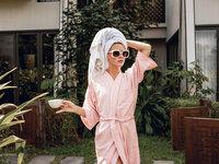 Woman wearing a pink robe, sunglasses, and a towel wrapped around her hair, holding a teacup while standing outdoors near a lush garden and modern building.