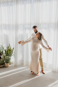 photo of pregnant woman holding husbands hands while they stand in front of large windows taken by Lifestyle photographer Madison WI