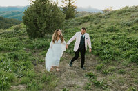 Wedding and elopement photographer located in Rexburg Idaho, Adriana Montoya Photo. A groom leads his bride by the hand, walking towards the camera. They are walking up a small hill with other hills and trees behind them.