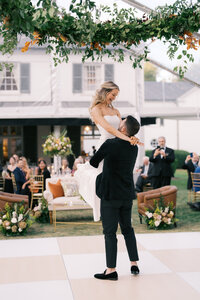 Maddie and Garrett share a joyful first dance at their wedding at Boxwood Estate. Garrett lifts Maddie into the air as they both smile, surrounded by soft lighting and elegant decor. Maddie wears a stunning white wedding gown, and Garrett is dressed in a sharp suit. The romantic atmosphere of the venue, with its beautiful chandeliers and glowing ambiance, enhances this special, carefree moment between the newlyweds.