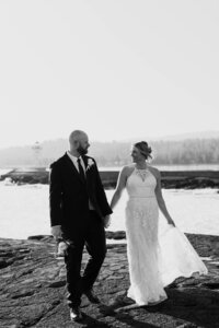 A newlywed couple holding hands and walking along a shoreline 