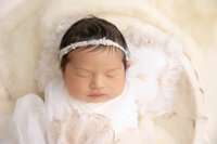 newborn baby girl sleeping sleeping in a decorative wooden bowl and wrapped with a soft buttery cream blanket and wearing a cream-flowered headband.