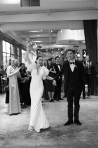 A newlywed couple holding hands and celebrating with their guests cheering behind