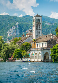 A scenic view of Lake Orta with a historical building featuring arched windows by the water's edge. A tall bell tower rises behind it, surrounded by lush greenery and hills under a partly cloudy blue sky.
