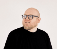 Bearded man with black-rimmed glasses and a minimalist style, dressed in a black shirt and gazing off-camera with a neutral white backdrop.