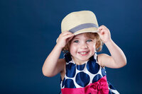 Three year old girl with a big smile holding onto a hat on her head.