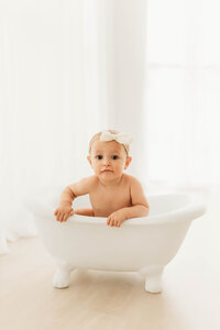 baby girl in a white studio sitting in a white clawfoot baby tub prop