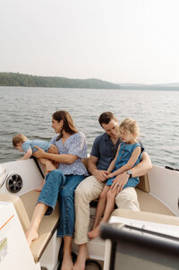A family of four cozies up on a boat drifting across a calm lake. Both parents hold their children close — the mother cradles their baby boy as he leans curiously over the edge, while the father lifts their smiling daughter in his arms. Dressed in soft knits and lake-weather layers, they glow in the golden light. Captured with the finesse of a New England photographer, the image radiates fine art elegance and a tender, documentary-style warmth.