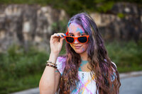 Fun and colorful senior picture of a wavy haired girl wearing orange sunglasses, and covered in different colors of Holi Powder. Captured by Springfield, MO photographer Dynae Levingston.