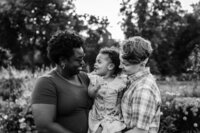 A black and white photo of a happy family of three, standing closely together outdoors. The parents, each holding one side of their toddler, are smiling at the child, who looks back at them with a joyful expression.