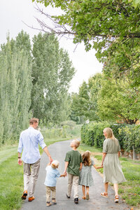 Two parents and their three children all hold hands while walking down a gravel path, lush green trees surrounding them. Taken by Cassie Lee Photography