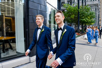 two grooms hold hands in navy blue suits as they walk